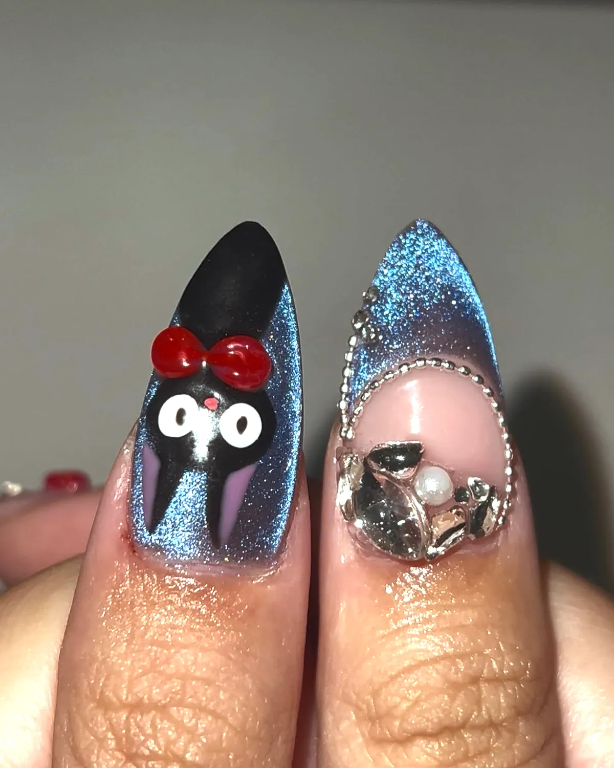 Jiji the black cat with a red bow on a blue cat-eye nail, next to a nail with crystals and a pearl